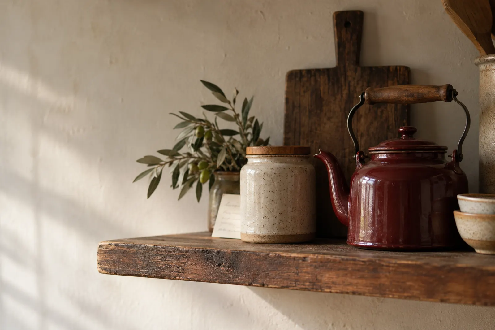 A rustic kitchen shelf in warm light: an olive branch in a jar, a stoneware canister, a deep-red kettle, and a handwritten card.